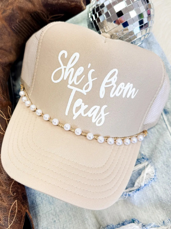 Beige trucker hat with 'She's from Texas' text and pearl embellishments on a denim background.