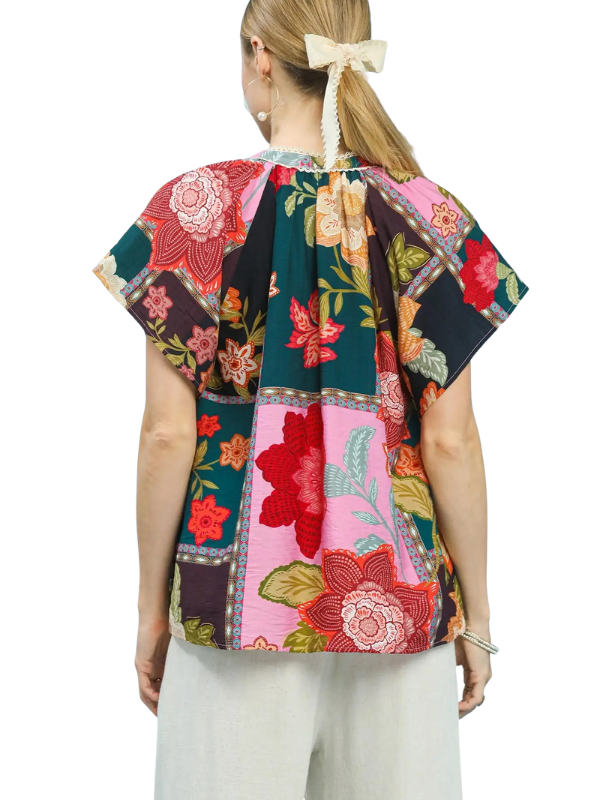 A woman shown from the back wearing a short-sleeve patchwork floral top with a cream hair bow, set against a plain white background.