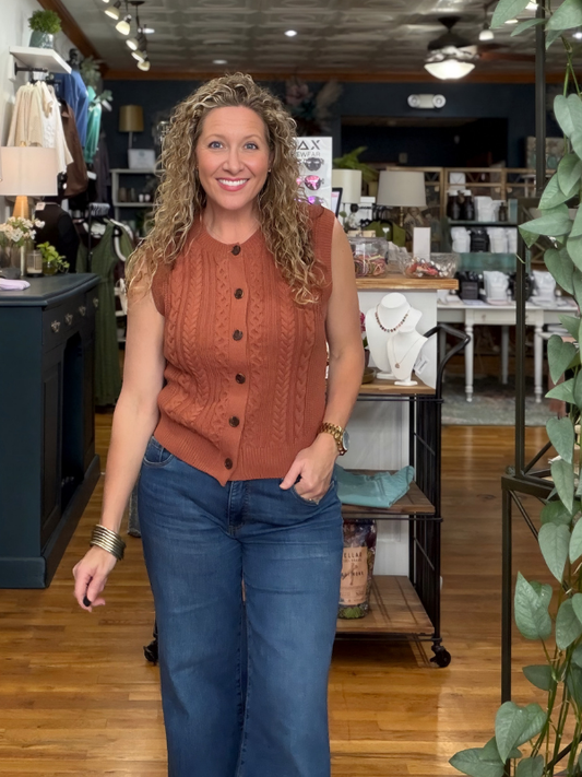 A woman with curly blonde hair stands in a boutique wearing a sleeveless rust-orange cable-knit button-front vest and blue jeans.