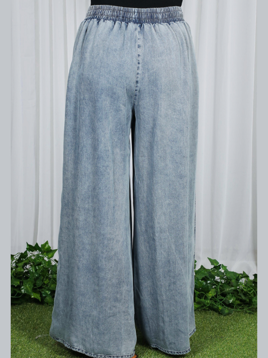 Light blue denim wide-leg pants on a white background with green plants at the base.
