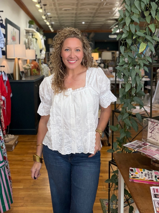 Woman in a white blouse and blue jeans standing in a store with plants and shelves in the background.