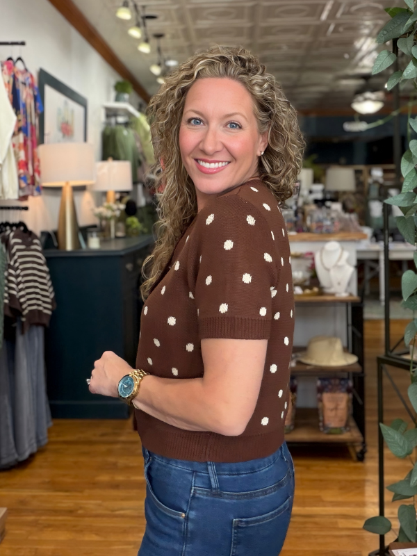 A smiling woman with curly blonde hair stands in a boutique shop, wearing a brown short-sleeve knit sweater with white polka dots and blue jeans.