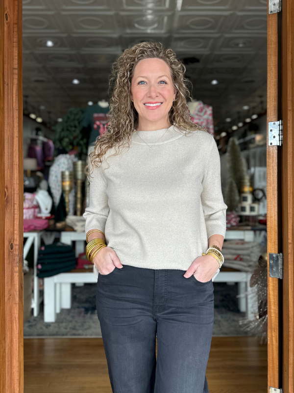 Woman standing in a store doorway wearing a light sweater and dark pants.