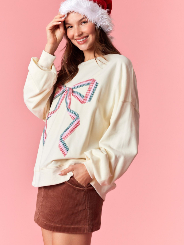 Woman wearing a cream sweater with a large bow design and a Santa hat on a pink background