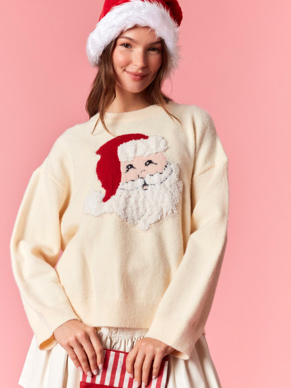 Person wearing a cream sweater with a Santa Claus design and a red Santa hat on a pink background