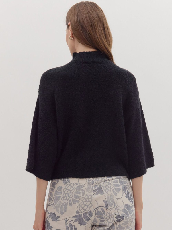 Person wearing a black poncho over patterned pants on a plain background