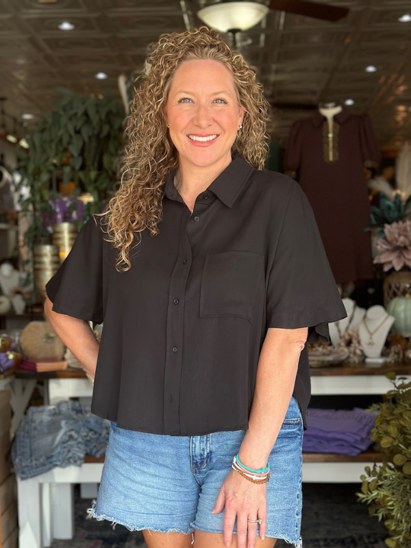 Woman wearing a black shirt and denim shorts standing in a store with various items in the background.