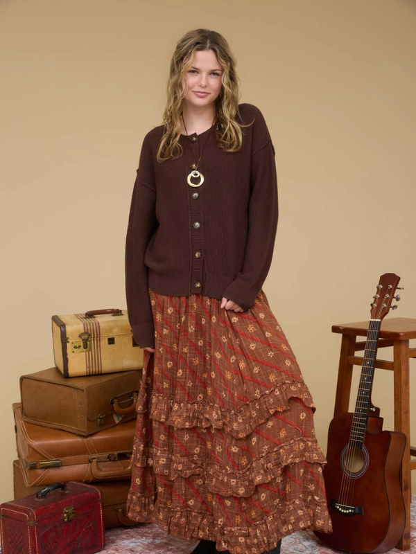 Woman wearing a brown cardigan and patterned skirt standing next to vintage suitcases and a guitar.