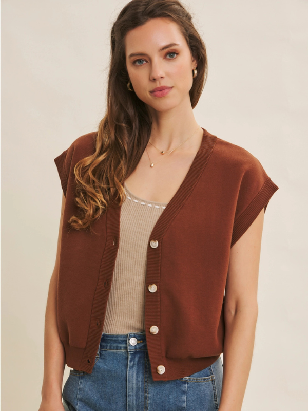 Woman wearing a brown vest over a beige top and blue jeans on a plain background