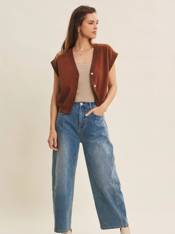Woman wearing a brown vest over a beige top and blue jeans on a plain background