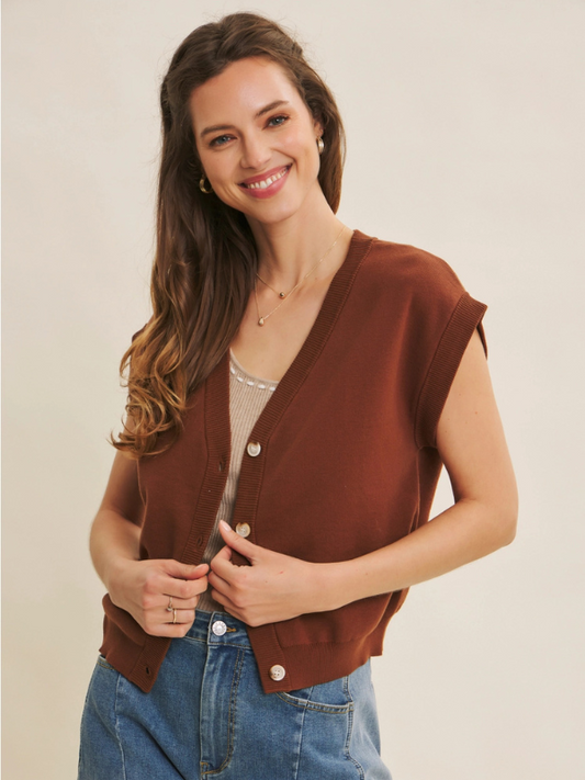 Woman wearing a brown sleeveless cardigan over a beige top and blue jeans on a beige background