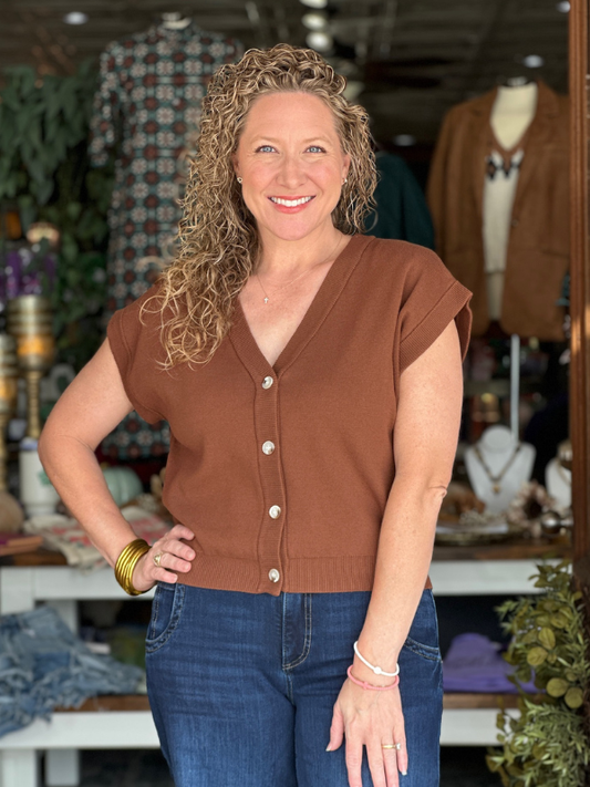Woman wearing a brown top and blue jeans standing in a store with clothing and decor in the background.