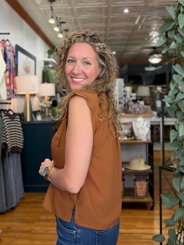 A woman with curly blonde hair wearing a sleeveless rust-brown top and blue jeans, smiling and looking back in a boutique setting.