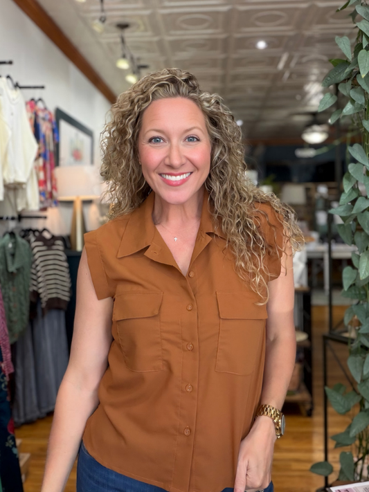 A smiling woman with curly blonde hair wearing a sleeveless brown button-up shirt and blue jeans, standing inside a clothing store.
