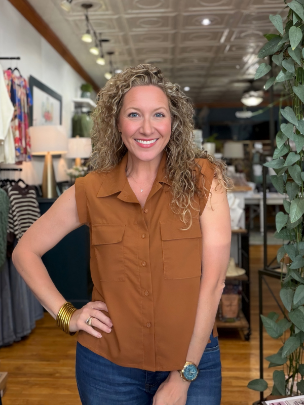 A smiling woman with curly blonde hair stands in a boutique wearing a sleeveless brown button-front collared shirt and blue jeans.