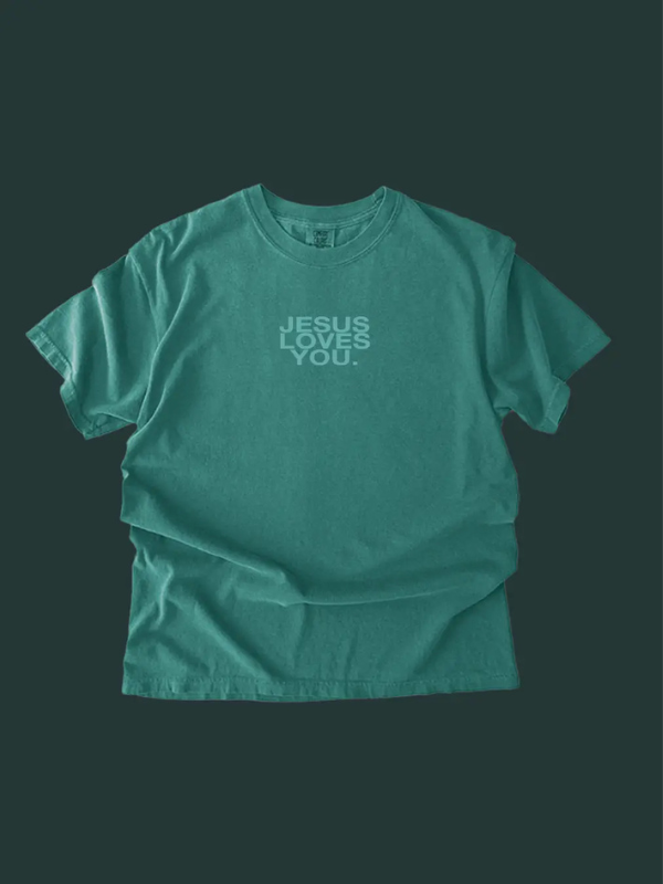 A teal green short-sleeve t-shirt with the text 'JESUS LOVES YOU.' printed in a lighter green font on the chest, displayed against a dark green background.