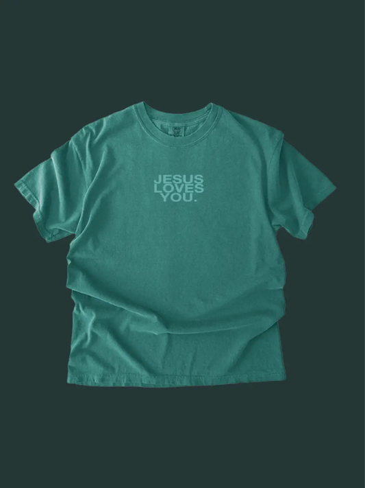 A teal green short-sleeve t-shirt with the text 'JESUS LOVES YOU.' printed in a lighter green font on the chest, displayed against a dark green background.