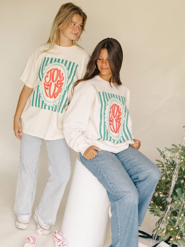 Two people wearing matching holiday-themed sweatshirts with a 'JOY' monogram design, sitting in front of a Christmas tree.