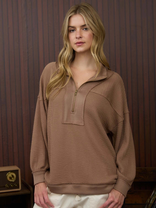 Woman wearing a brown ribbed sweater with a zipper against a wooden paneled wall.