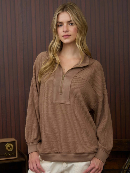 Woman wearing a brown ribbed sweater with a zipper against a wooden paneled wall.