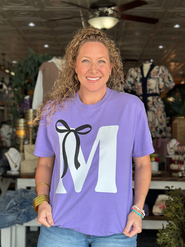 Woman wearing a purple t-shirt with a large white letter 'M' and black bow design, standing in a store.