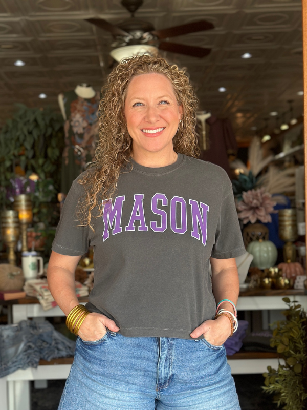 Woman wearing a gray t-shirt with 'MASON' printed in purple, standing in a store.