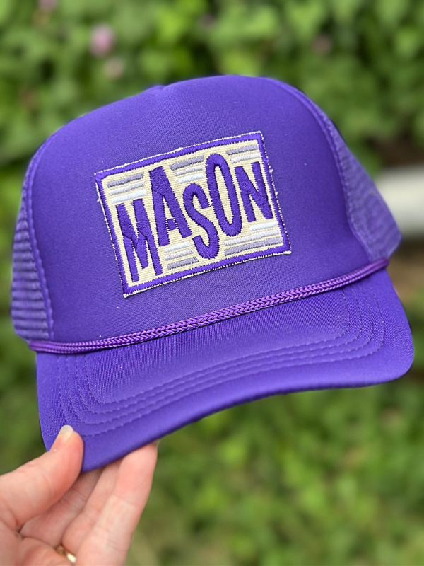 Purple trucker hat with 'MASON' patch held against a blurred green background
