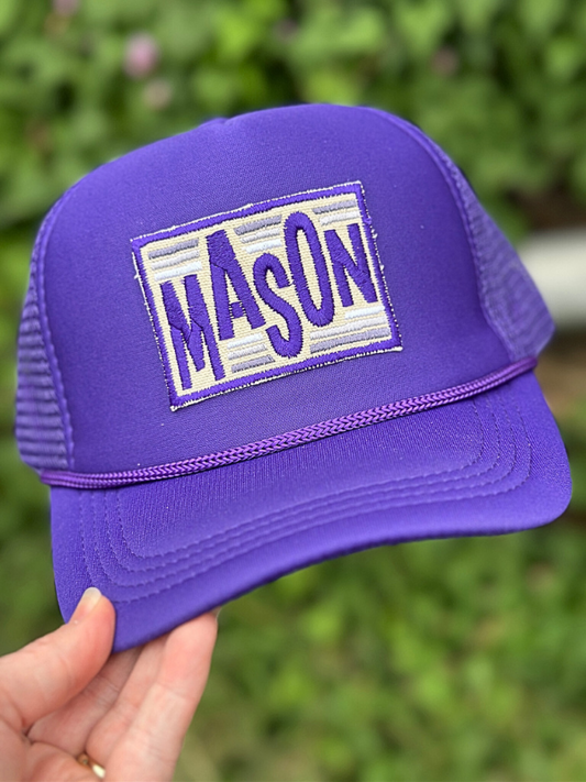 Purple trucker hat with 'MASON' patch held against a blurred green background
