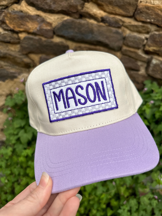 Lavender and beige cap with 'MASON' patch held in front of a stone wall.