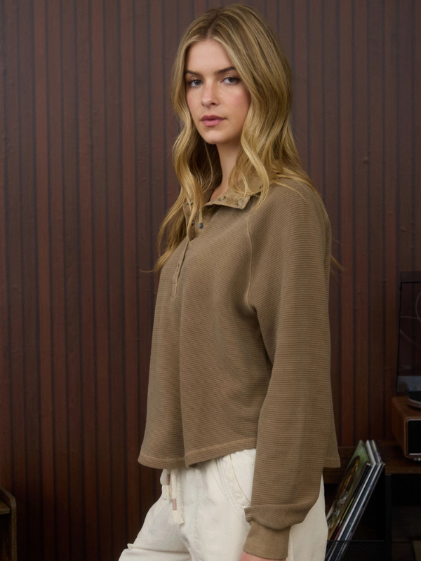 Woman wearing a brown sweater and white pants against a wooden paneled wall.