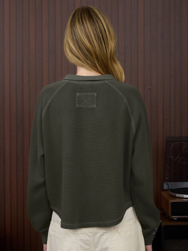 Person wearing a dark sweater with a small patch on the back against a wooden panel background.