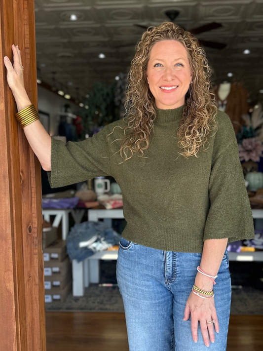 Woman in a green sweater and blue jeans standing indoors, smiling.
