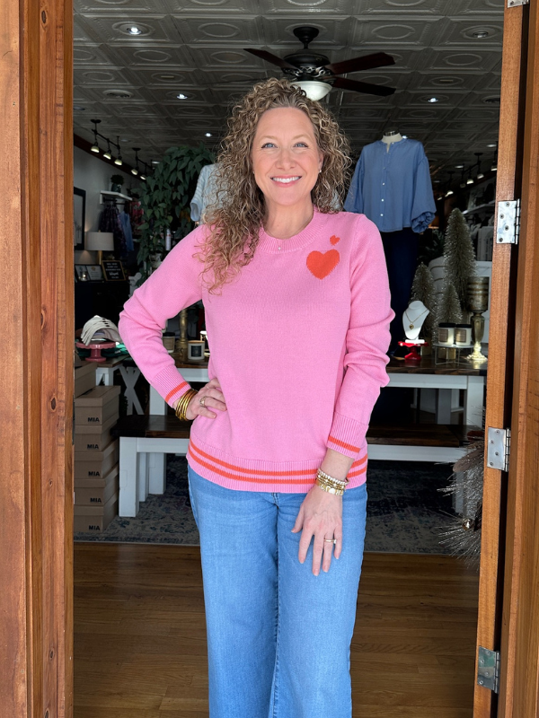 Woman wearing a pink sweater with a heart design and blue jeans standing in a store.