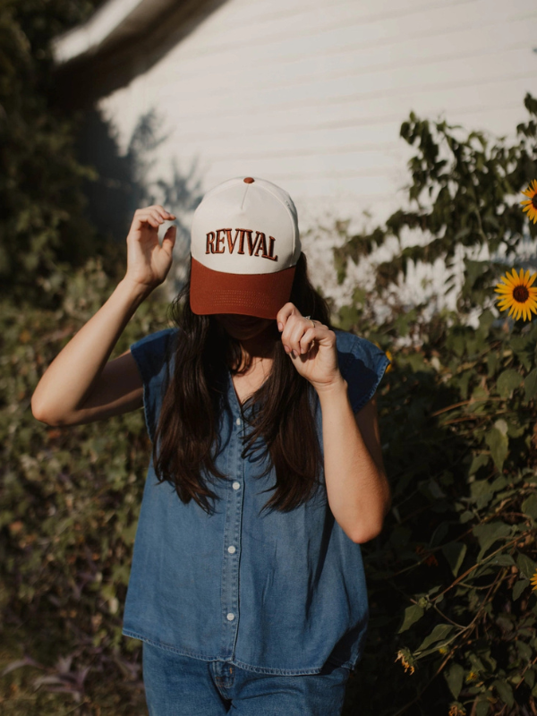 Person wearing a 'Revival' cap outdoors with greenery and sunflowers in the background