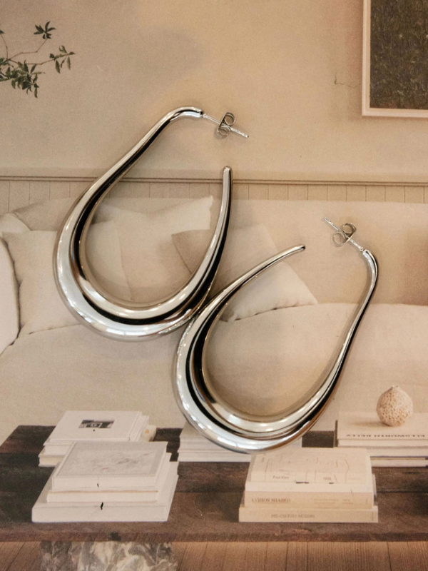 Large silver earrings displayed on a beige sofa with books and a plant in the background