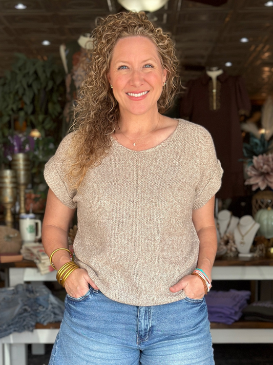 Woman wearing a beige sweater and blue jeans standing in a store with various items in the background.