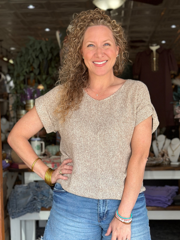 Woman wearing a beige knit top and blue jeans in an indoor setting with shelves and items in the background.