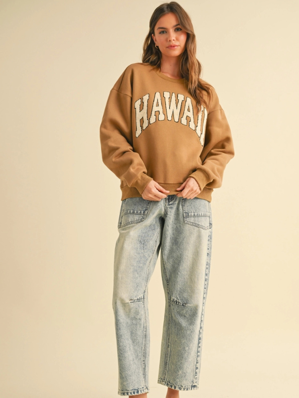 Woman wearing a brown 'HAWAII' sweatshirt and light blue jeans on a beige background