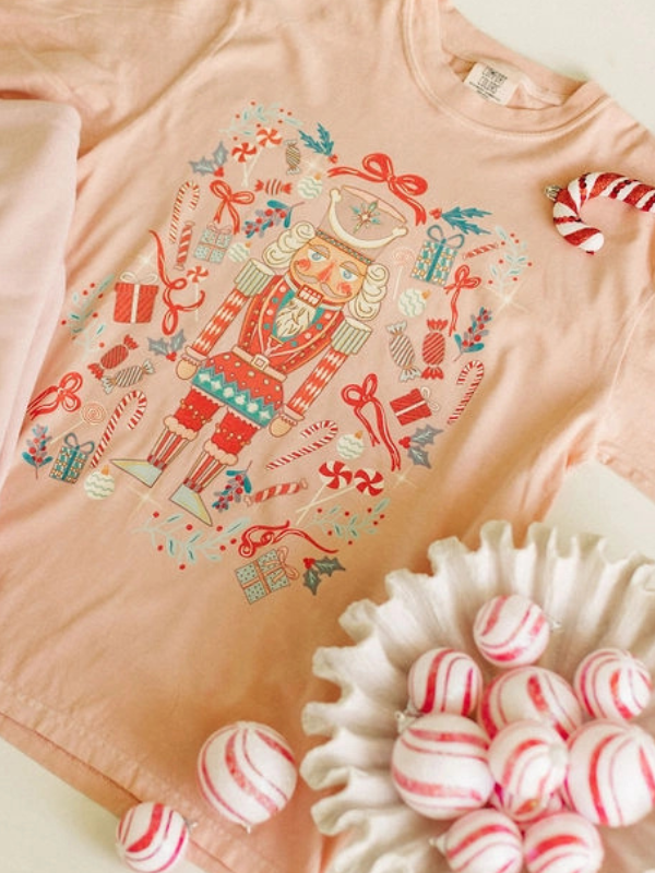 Pink t-shirt with nutcracker design and candy-themed elements, next to candy canes and a bowl of candy.