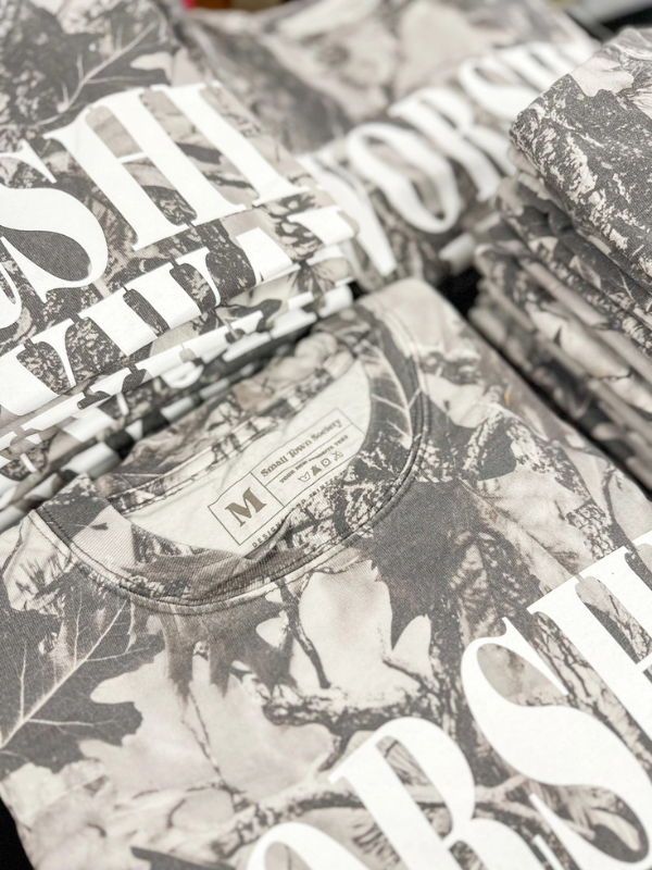 Stack of camouflage-patterned shirts with a brand logo.