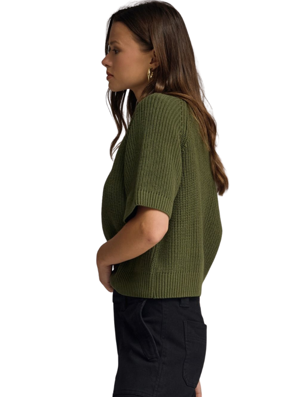 A woman shown in side profile wearing a short-sleeve olive green ribbed knit top and black pants against a plain white background.