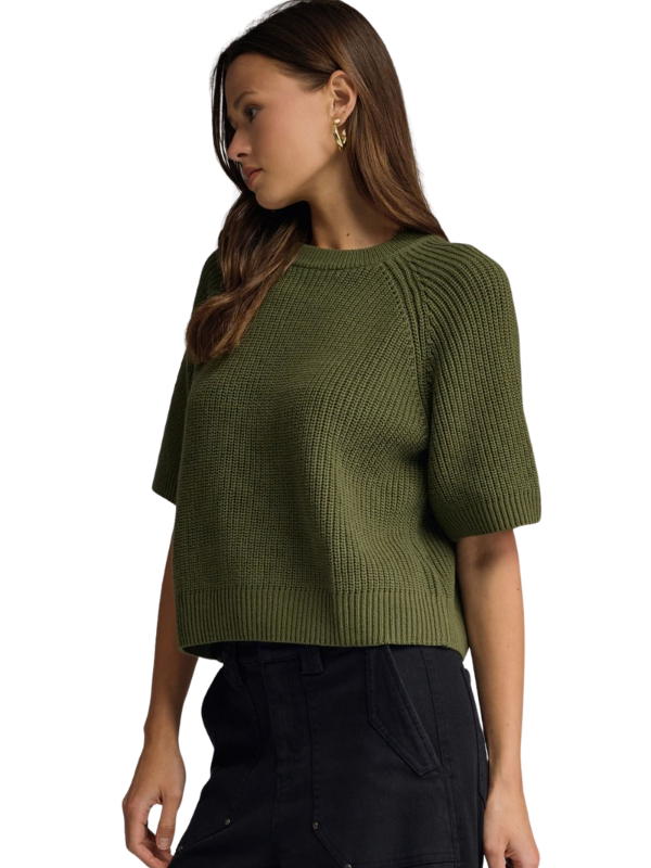 A woman wearing a textured olive green short-sleeve knit top and black pants, shown from the side against a plain white background.