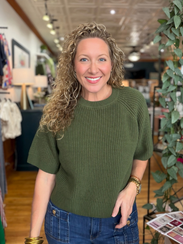 A smiling woman with curly blonde hair wearing an olive green short-sleeve ribbed knit sweater and blue jeans, standing inside a retail store.
