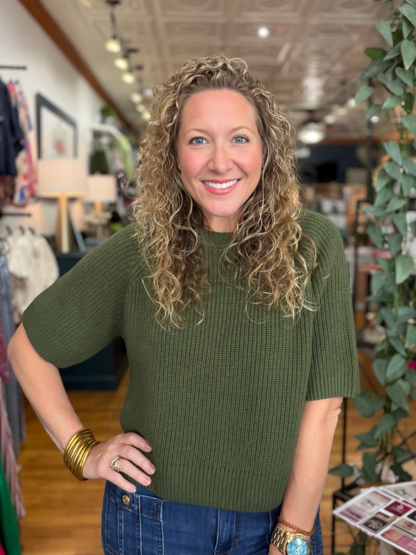 A smiling woman with curly blonde hair wearing a ribbed short-sleeve olive green knit top and blue jeans, standing in a boutique setting.