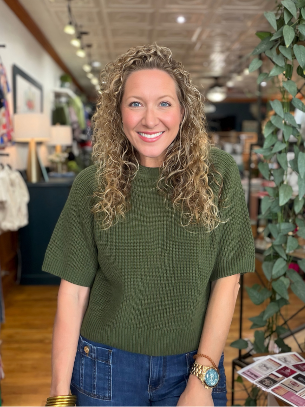 A smiling woman with curly blonde hair wearing a ribbed short-sleeve olive green top and blue jeans, standing inside a boutique store with plants and clothing racks in the background.