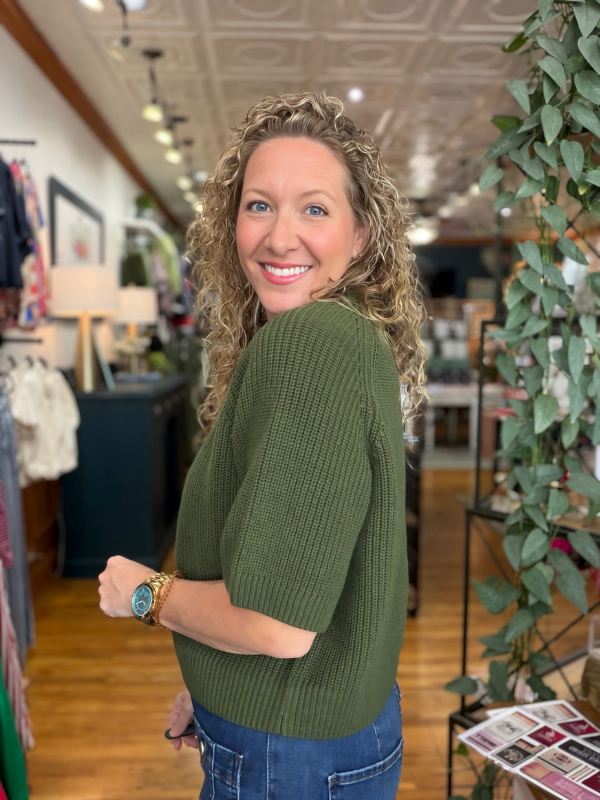 A smiling woman with curly blonde hair wearing an olive green ribbed knit sweater and blue jeans, standing inside a boutique store.