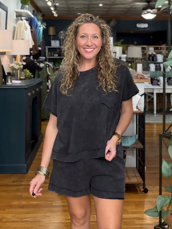 A smiling woman with curly blonde hair stands in a boutique wearing a matching black washed short-sleeve t-shirt and shorts set. The background shows a cozy, styled interior with wooden floors, shelves, and home decor.