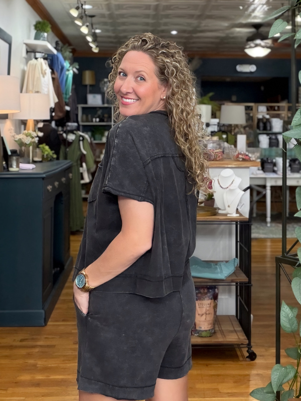 A woman with curly blonde hair wearing a dark gray washed short-sleeve top and matching shorts, standing in a boutique store.