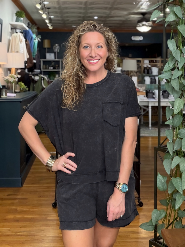 A woman with curly blonde hair stands in a boutique wearing a matching black washed short-sleeve t-shirt and shorts set. The background shows a retail store interior with wooden floors, shelves, and plants.