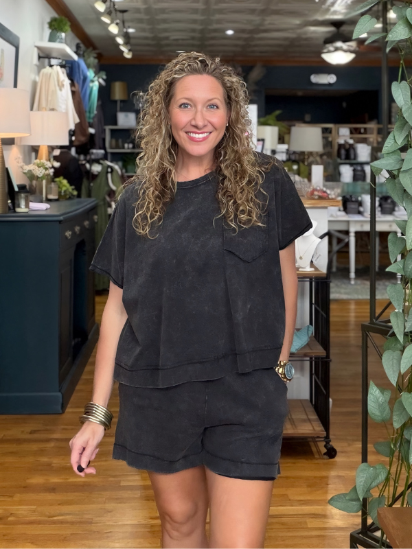 A woman with curly blonde hair stands in a boutique wearing a matching black washed short-sleeve t-shirt and shorts set. The background shows a retail store interior with wooden floors, shelves, and plants.